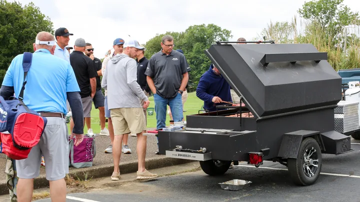 Golfers gathering beside an outdoor barbecue trailer.