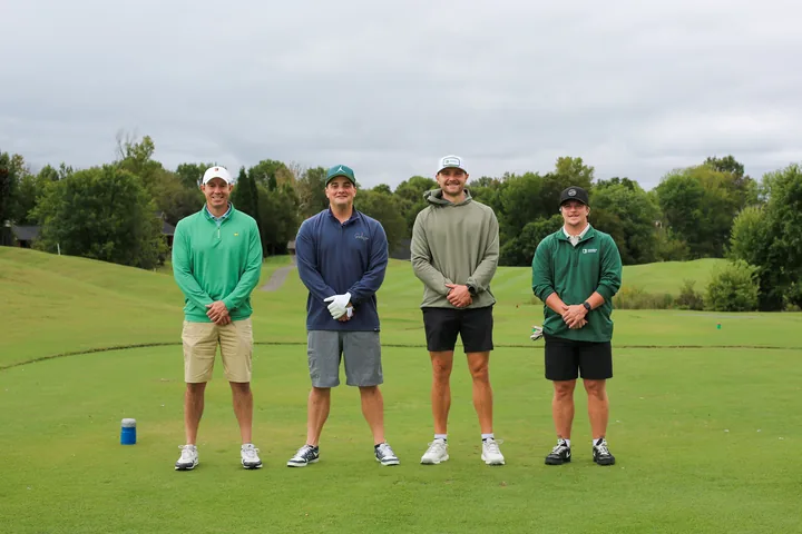 A golf foursome in green, navy, and gray shirts holding their clubs.