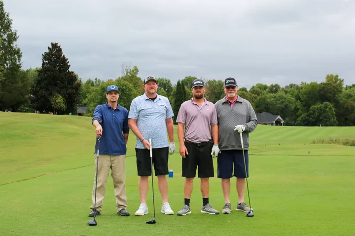 Four golfers in navy, blue, pink, and gray shirts posing with their clubs.