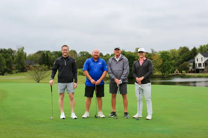 Four golfers in black, blue, and gray shirts posing near the water.