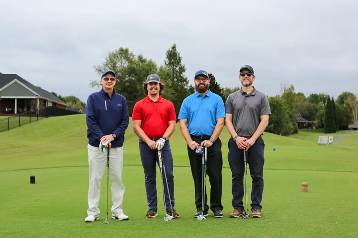 Four golfers in navy, red, blue, and gray shirts posing with their clubs.