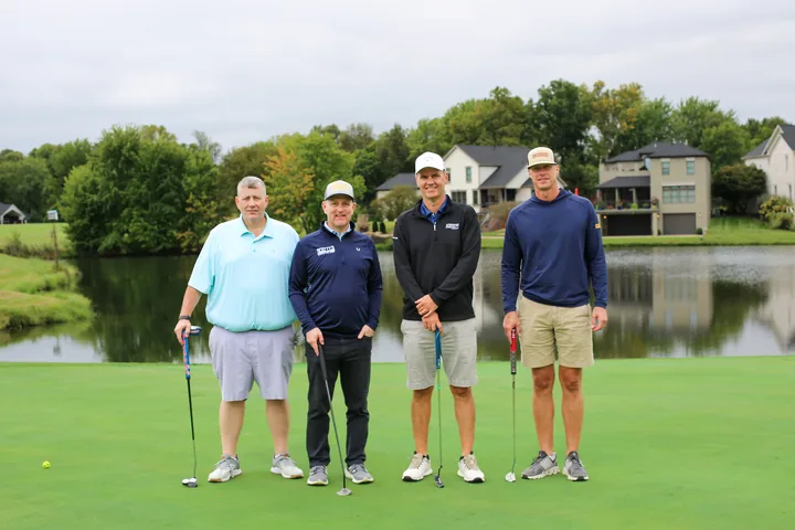 A golf foursome in light blue and navy shirts posing on the green.