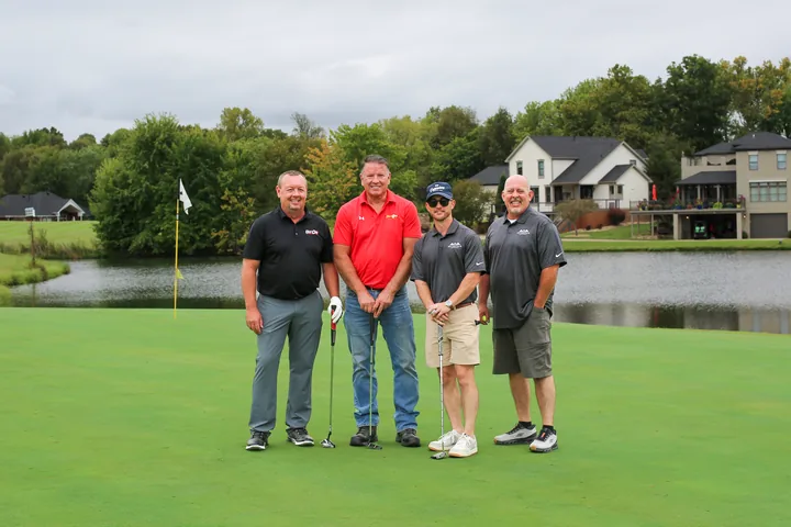 Four golfers in black, red, and gray shirts posing on the lakeside green.