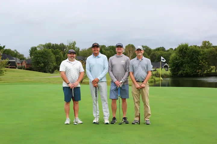 A golf foursome in white and gray shirts holding their clubs on the green.