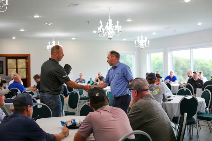 Two participants greeting each other as guests gather at tables in the clubhouse.